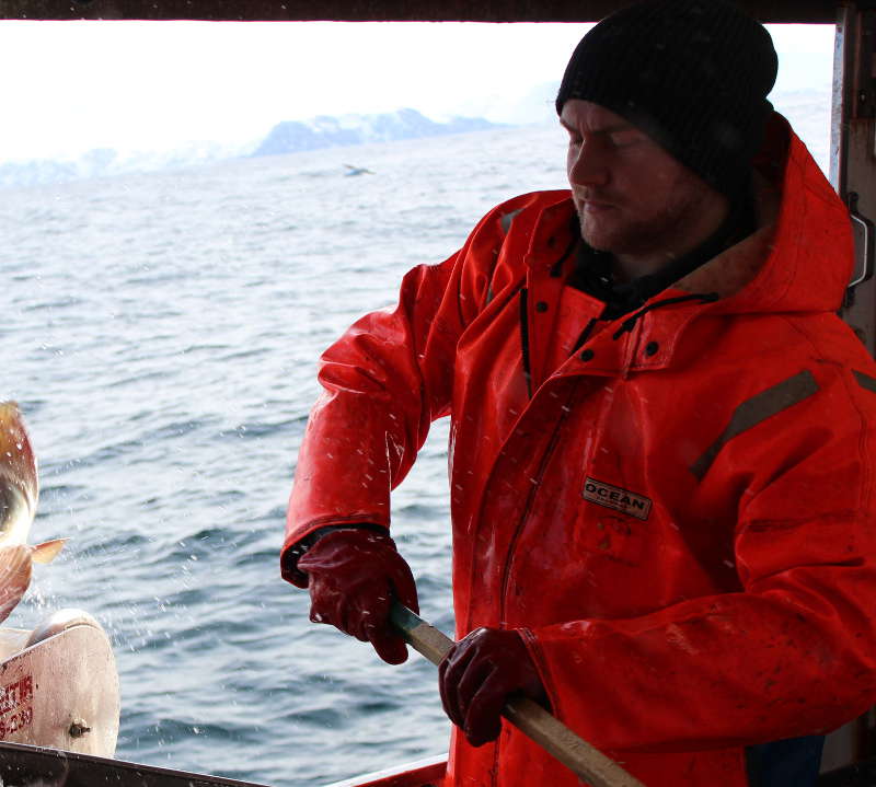 Fisker i oransje jakke på dekk, med hav og fjell i bakgrunnen